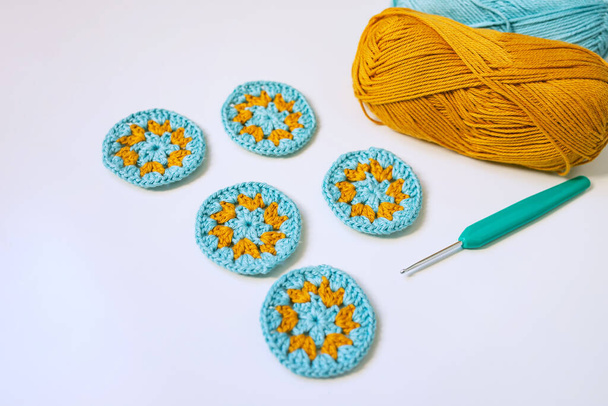 Side view of five round crochet motifs in turosque and yellow colors, a hook and skeins of yarn on smooth white surface. Crocheting round motifs concept. Selected focus. - Photo, Image