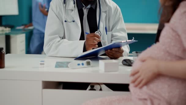 Afrikanisch-amerikanischer Arzt macht sich Notizen in Absprache mit Schwangeren und schreibt einen Kontrollbericht über Dokumente. Frau mit Schwangerschaftsbauch bittet männlichen Geburtshelfer um Mutterschaftsberatung. - Filmmaterial, Video