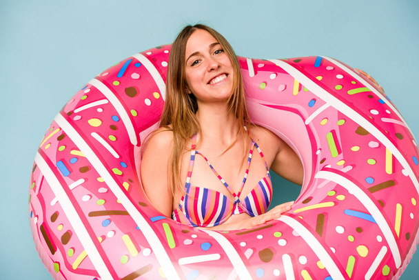 Young caucasian woman holding air mattress isolated on blue background - 写真・画像