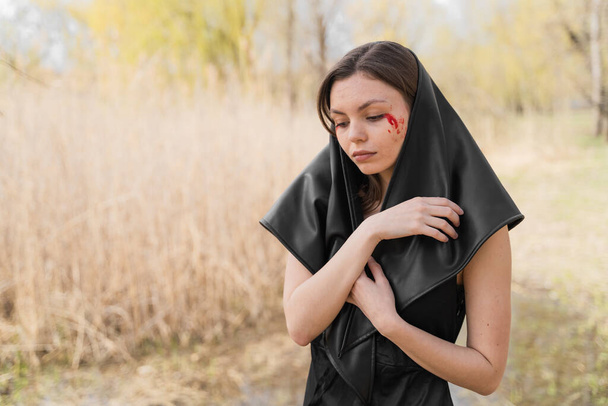 Young widow in a black mourning dress and headscarf suffering, praying and crying with bloody tears. Ukrainian woman. Victim of war. Stop war, fighting and genocide in Ukraine. - Foto, afbeelding