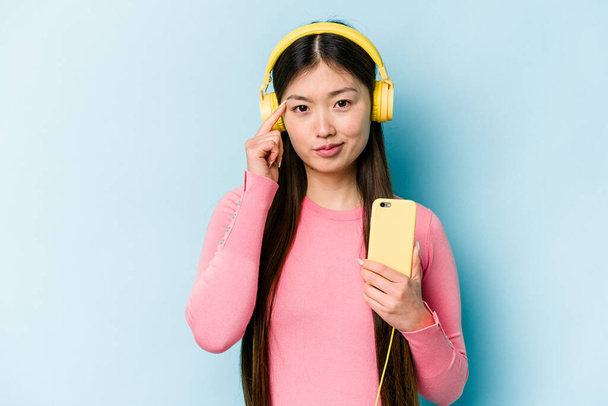Joven mujer asiática escuchando música aislada sobre fondo azul apuntando templo con el dedo, pensando, enfocado en una tarea. - Foto, imagen