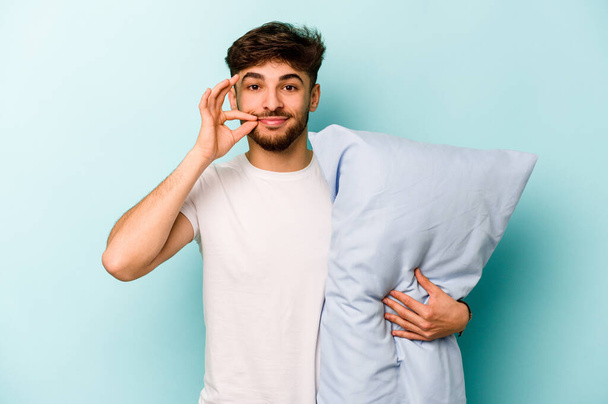 Молодой латиноамериканец, одетый в пижаму, держит подушку, изолированную на синем фоне, с пальцами на губах, хранящими секрет.. - Фото, изображение