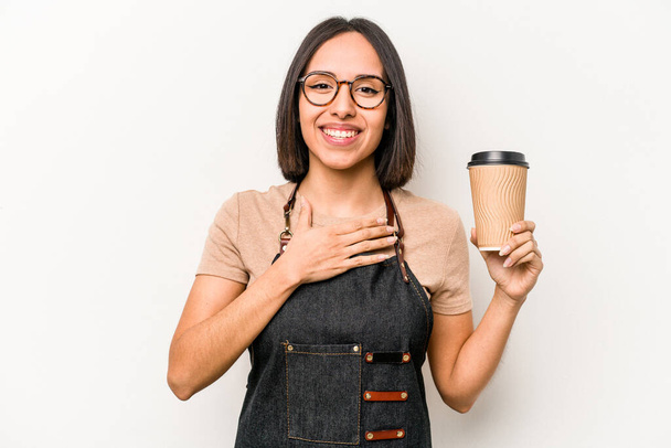 Молодая кавказская официантка, держащая в руках кофе, изолированный на белом фоне, громко смеется, держа руку на груди. - Фото, изображение