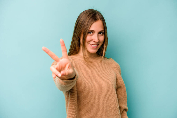 Young caucasian woman isolated on blue background joyful and carefree showing a peace symbol with fingers. - Foto, Imagen