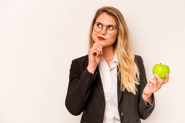 Fiatal üzleti kaukázusi nő kezében egy alma elszigetelt fehér háttér néz oldalra kétes és szkeptikus kifejezés. - Fotó, kép