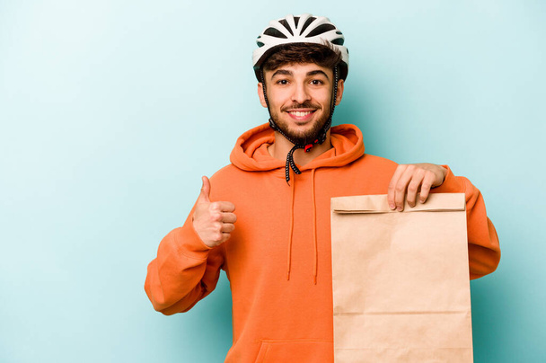 Молодой латиноамериканец, одетый в мотоцикл со шлемом, держит еду, изолированную на синем фоне, улыбаясь и поднимая большой палец вверх - Фото, изображение