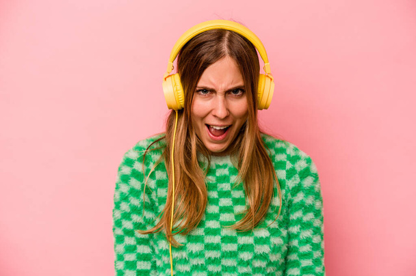 jonge Kaukasische vrouw luisteren naar muziek geïsoleerd op roze achtergrond schreeuwen zeer boos en agressief. - Foto, afbeelding
