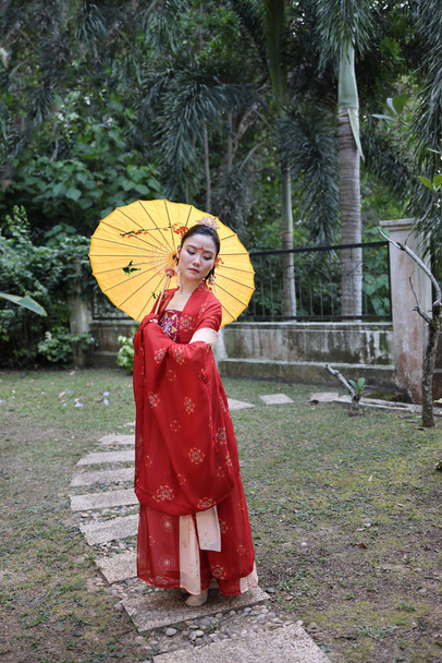 Młoda atrakcyjna azjatycka kobieta nosi tadeonal chiński czerwony hanfu długa spódnica sukienka kostium szalik spinka do włosów ucho urządzone parasol odkryty zielony ogród patio - Zdjęcie, obraz