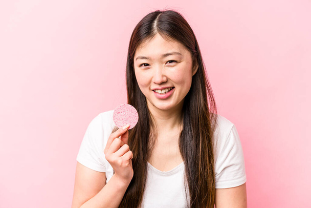 若いですアジアの女性を保持顔スポンジ隔離されたピンクの背景笑いと楽しみを持って. - 写真・画像