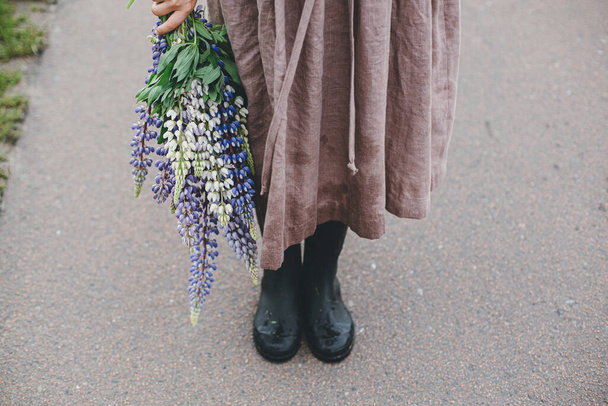 Close up of woman in rustic dress and gumboots holding lupine bouquet in summer countryside. Cottagecore aesthetics. Young female with wildflowers after rain on rural road.Moody image - Foto, imagen