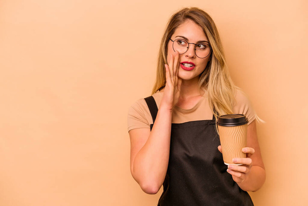 Restaurantkellner kaukasische Frau hält einen Kaffee zum Mitnehmen isoliert auf beigem Hintergrund sagt eine geheime heiße Bremse Nachrichten und Blick zur Seite - Foto, Bild
