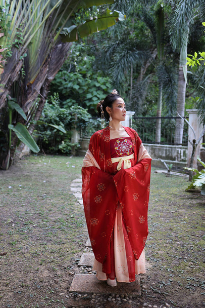 Νεαρή ελκυστική Ασιάτισσα γυναίκα φορώντας tadeonal κινεζική κόκκινο hanfu μακριά φούστα φόρεμα κοστούμι φουλάρι φουρκέτα φορώντας υπαίθριο πράσινο κήπο - Φωτογραφία, εικόνα