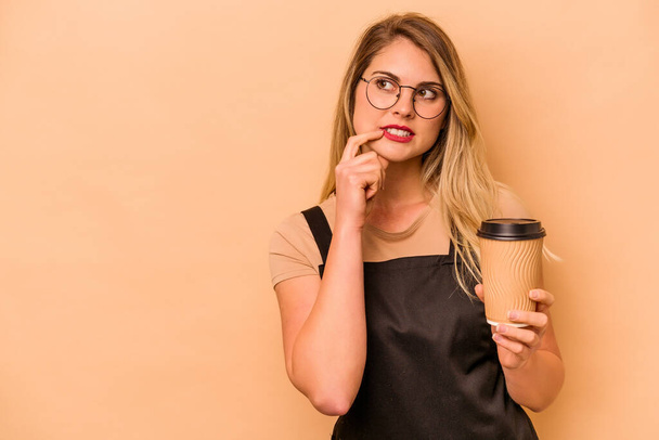 Restaurantkellner kaukasische Frau hält einen Kaffee zum Mitnehmen isoliert auf beigem Hintergrund und denkt entspannt über etwas nach, das einen Kopierraum betrachtet. - Foto, Bild