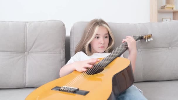 Triste petite fille jouant de la guitare, elle se repose assise sur le canapé à la maison. Jeune musicienne. Vidéo 4k - Séquence, vidéo