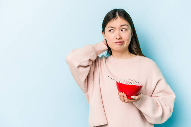 Joven mujer asiática sosteniendo un tazón de cereales aislado sobre fondo azul tocando la parte posterior de la cabeza, pensando y haciendo una elección. - Foto, imagen