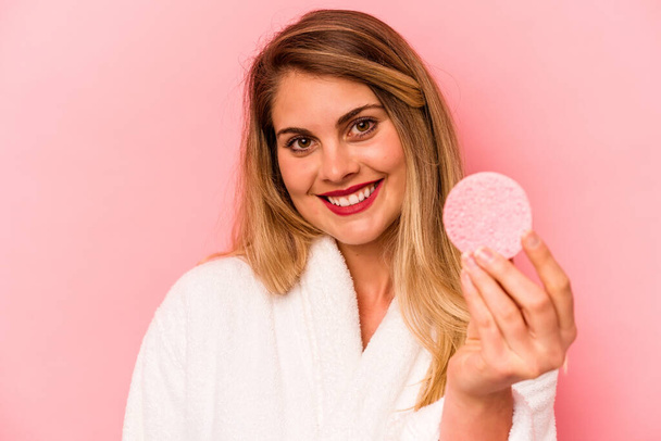 Young caucasian woman holding facial sponge isolated on pink background - Foto, afbeelding