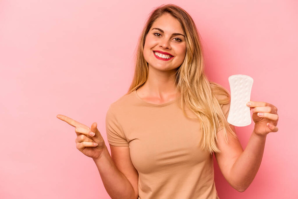 Young caucasian woman holding sanitary napkin isolated on pink background smiling and pointing aside, showing something at blank space. - Foto, immagini