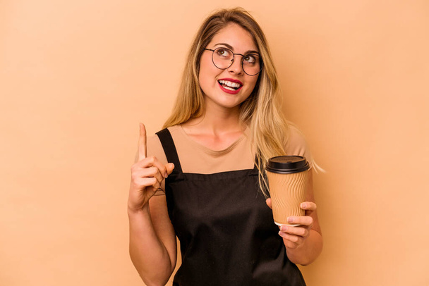 Restaurantkellner kaukasische Frau mit einem Kaffee zum Mitnehmen isoliert auf beigem Hintergrund zeigt auf den Kopf mit geöffnetem Mund. - Foto, Bild