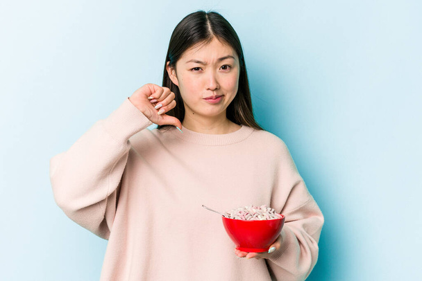 Giovane donna asiatica in possesso di una ciotola di cereali isolati su sfondo blu mostrando un gesto antipatico, pollici verso il basso. Concetto di disaccordo. - Foto, immagini