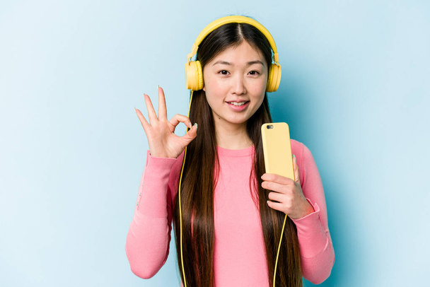 Junge asiatische Frau hört Musik isoliert auf blauem Hintergrund fröhlich und zuversichtlich zeigt ok Geste. - Foto, Bild