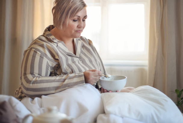 A happy overweight woman with headphones and smartphone having breakfast in bed at home. - Valokuva, kuva