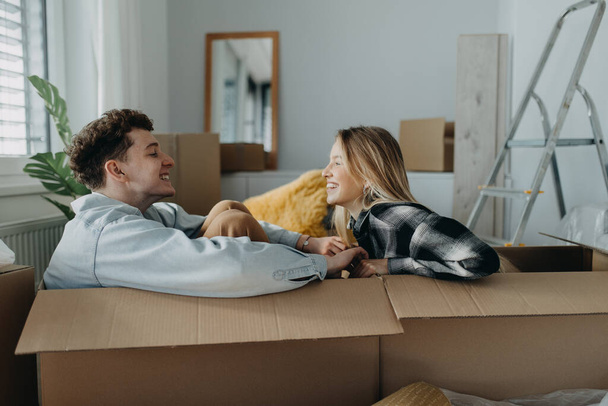 A cheerful young couple in their new apartment, sitting in boxes and having fun. Conception of moving. - Foto, immagini