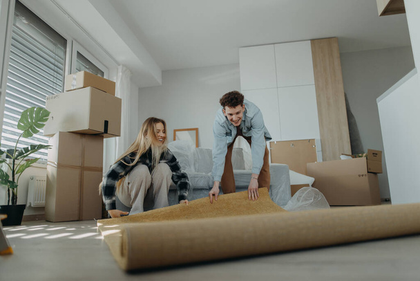A cheerful young couple in their new apartment, rolling out carpet. Conception of moving. - Фото, изображение
