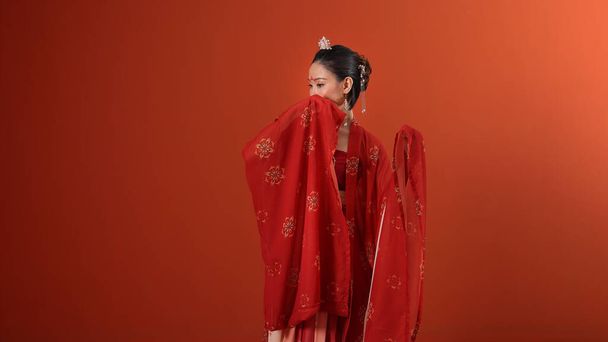 Jovem atraente mulher asiática vestindo tadeonal chinês vermelho hanfu vestido de saia longa cachecol hairpin brinco interior na parede de fundo leia marron - Foto, Imagem
