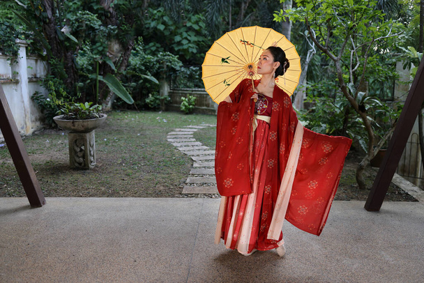Young attractive Asian woman wearing tadeonal Chinese red hanfu long skirt dress costume scarf hairpin earing decorated umbrella outdoor green garden patio - Photo, Image
