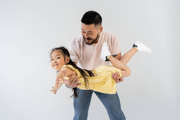 cheerful asian man holding in arms smiling daughter in yellow dress isolated on grey - Zdjęcie, obraz