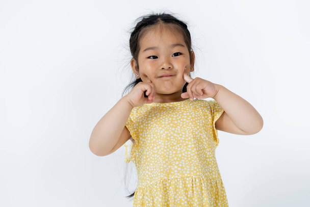 portrait of preschooler asian girl pointing at cheeks isolated on grey  - Fotografie, Obrázek