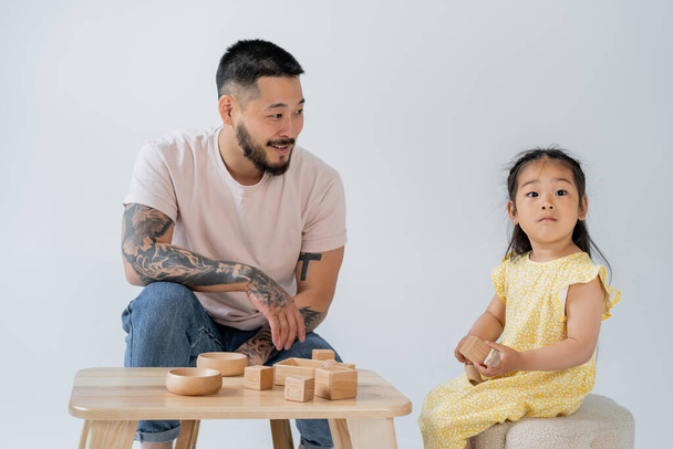 cheerful and tattooed asian man looking at brunette daughter playing with wooden toys isolated on grey - Фото, зображення