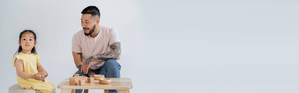 cheerful and tattooed asian man looking at brunette daughter playing with wooden toys isolated on grey, banner - Photo, Image