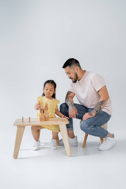 tattooed asian man looking at brunette daughter playing with wooden toys on grey - Photo, Image