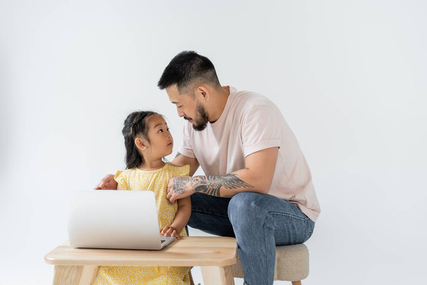 tattooed father looking at surprised asian daughter near laptop on wooden table isolated on grey - Fotó, kép