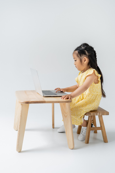 side view of asian girl in yellow dress using laptop on grey - Foto, afbeelding