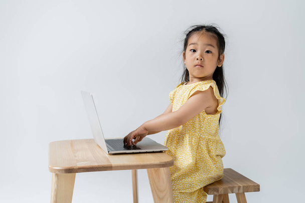 aziatische kleuter meisje in gele jurk met behulp van laptop en kijken naar camera geïsoleerd op grijs - Foto, afbeelding