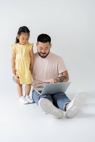 cheerful asian father using laptop near daughter in yellow dress on grey - Fotoğraf, Görsel