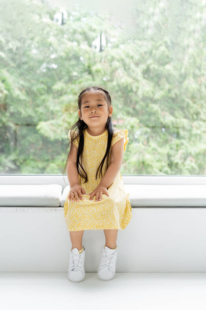 full length of cheerful asian girl in yellow dress sitting on soft pillow on windowsill  - 写真・画像