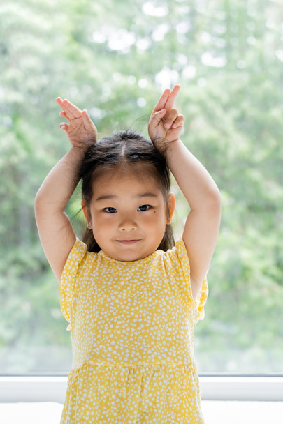 portrait of brunette and preschooler asian girl in yellow dress showing bunny ears near window at home - Zdjęcie, obraz