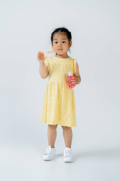 full length of asian girl in yellow dress holding bottle with soap bubbles on grey - 写真・画像