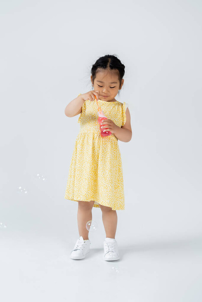 full length of asian preschooler child in yellow dress holding bottle with soap bubbles on grey - Foto, afbeelding