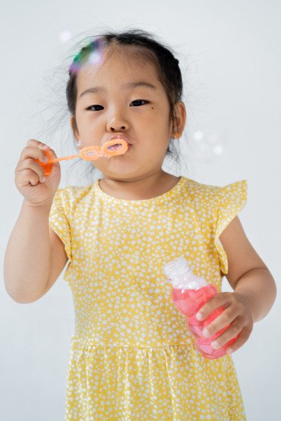 preschooler asian girl in dress holding bottle and blowing soap bubbles isolated on grey - Photo, Image