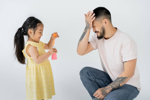 asian preschooler kid in yellow dress holding bottle with soap bubbles near tattooed father isolated on grey - Фото, зображення