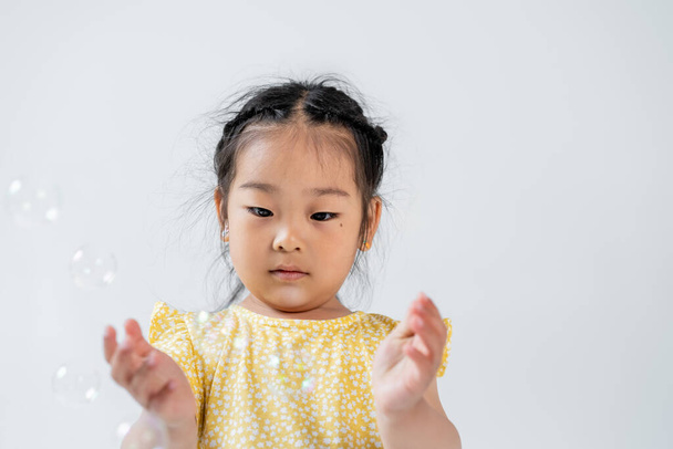 portrait of asian preschooler girl in yellow dress near blurred soap bubbles isolated on grey  - Foto, Imagem