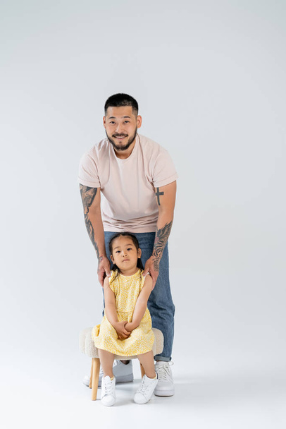 full length of smiling asian man with tattoos standing near preschooler daughter on grey - Foto, afbeelding