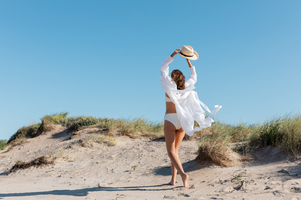 back view of barefoot young woman in white swimsuit and shirt walking with straw hat on sandy beach - Fotografie, Obrázek