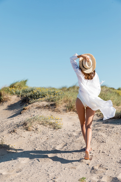 back view of barefoot young woman in white swimsuit and shirt walking with sun hat on sandy beach - Photo, Image