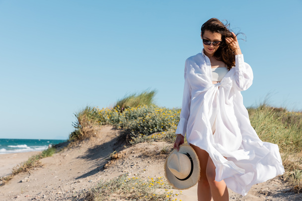 smiling young woman in sunglasses and white shirt holding straw hat on sandy beach - Φωτογραφία, εικόνα