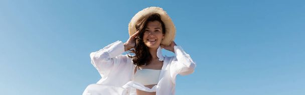 vista basso angolo di felice giovane donna in costume da bagno e camicia bianca indossando cappello di paglia contro il cielo blu, banner - Foto, immagini